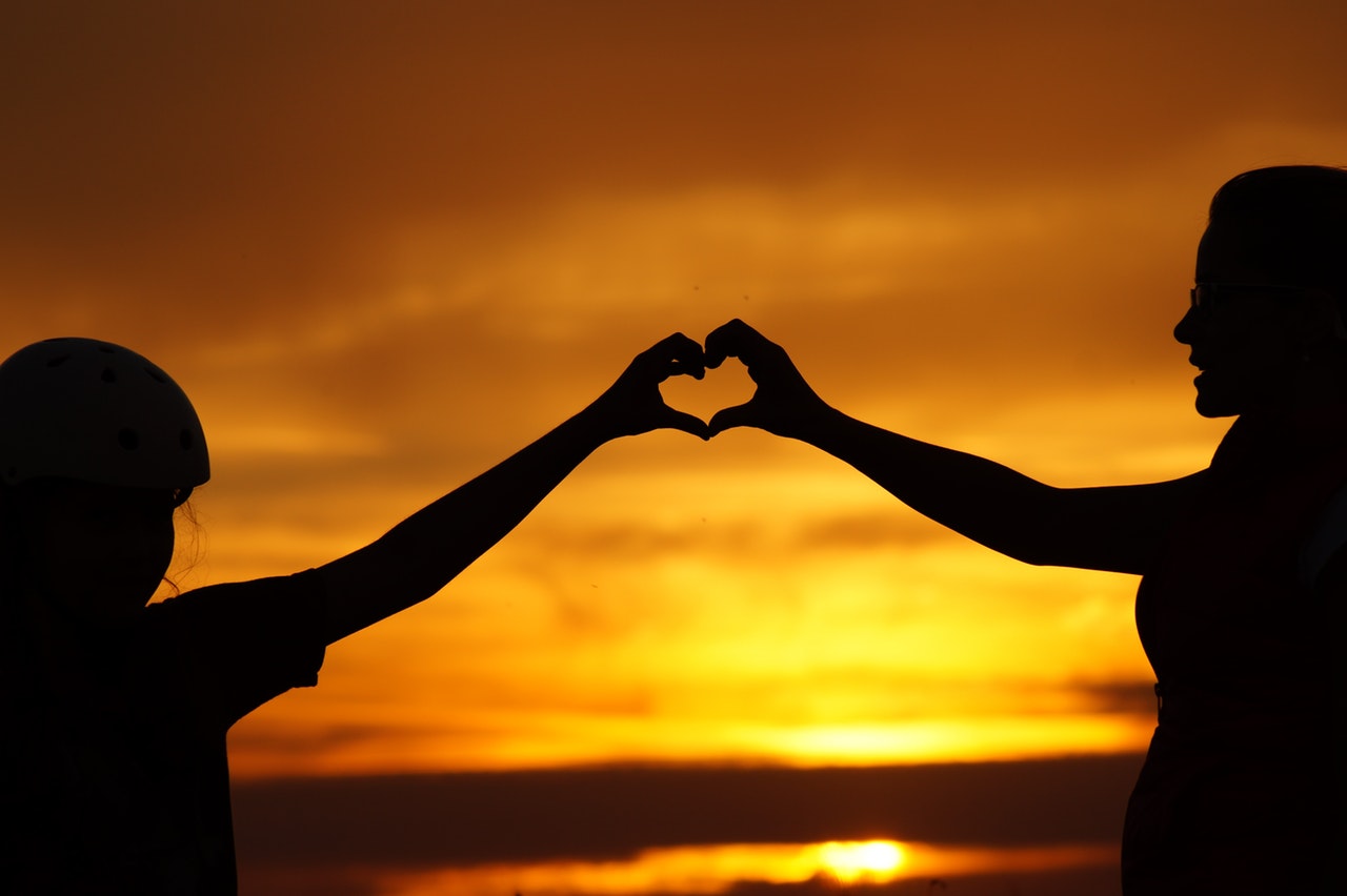 Two people making a heart shape with their hands, against the setting sun