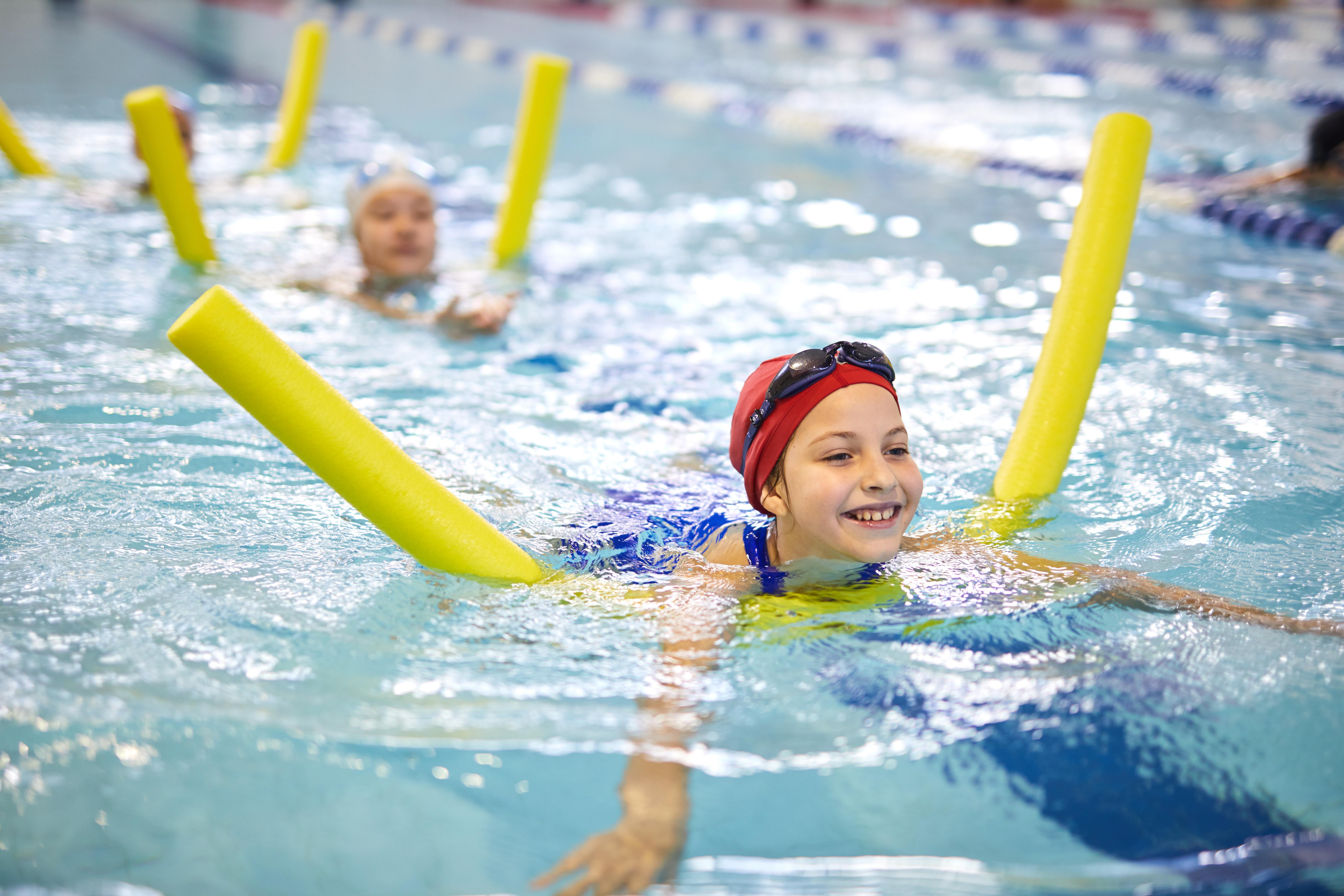 A young girl wearing a swimming hat and goggles, using a yellow float in a swimming pool